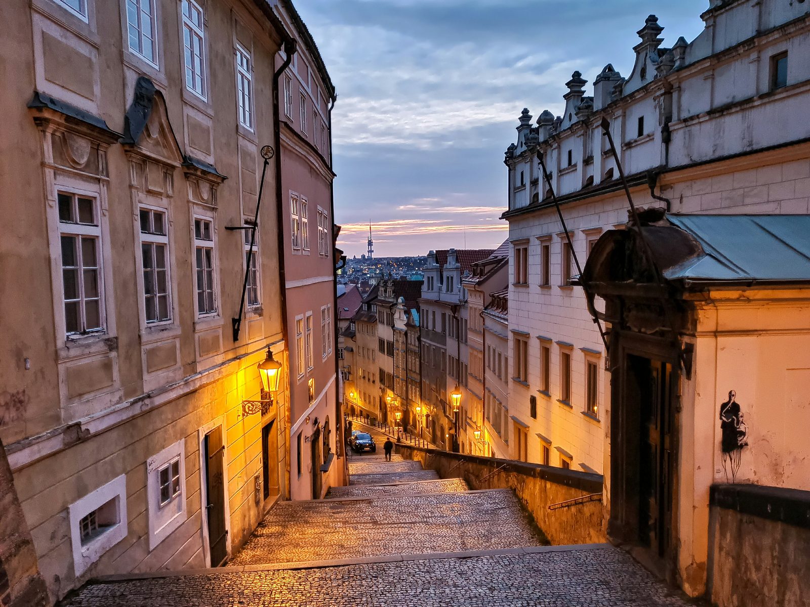 Rozsvícené plynové lampy nad Radnickými schody za soumraku, dole světla Malé Strany