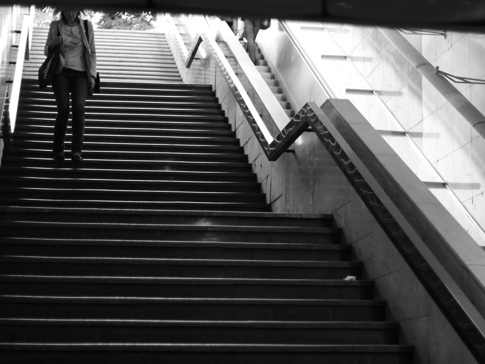 Schodiště v metru černobíle, postava sestupuje podél ostrých linií stupňů a zábradlí